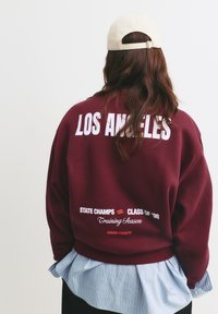 Bordeaux sweatshirt met witte tekst, met "LOS ANGELES" op de achterkant, gelaagd over een blauw gestreept shirt. De tekst bevat "STATE CHAMPS" en "HONOR SOCIETY."