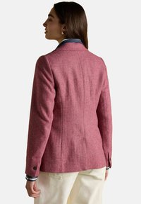 Žena oblečená v priliehavom ružovom sako s herringbone vzorom a gombíkovými manžetami, vrstvená na pruhovanej košeli, kombinovaná s svetlými nohavicami.