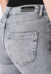 Grå denimjeans med en tettsittende design, med et svart merke ved midjen og en standard baklomme med sydd detalj.