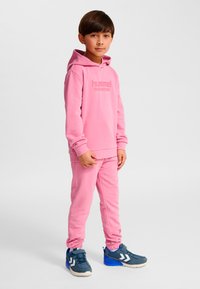 Rosa Kapuzenpullover und passende Jogginghose aus weichem Stoff, mit einem Logo auf der Vorderseite. Kombiniert mit blauen Sportschuhen.