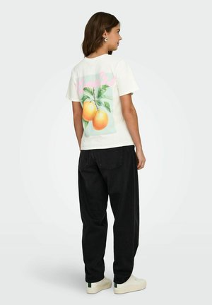 Femme debout de profil portant un t-shirt blanc avec un imprimé d'orange au dos, un pantalon ample noir et des baskets blanches.
