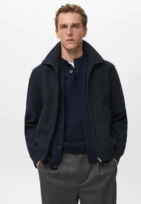 Giacca in misto lana navy con collo alto, chiusura con zip e texture morbida, abbinata a un maglione scuro e pantaloni grigi.
