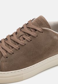 Sapatilha de camurça castanha com atacadores redondos, sola de borracha texturizada e uma discreta marcação em relevo na língua. Design limpo e minimalista.