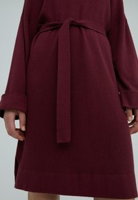 Bordeauxfarbenes geripptes Kleid mit tailliertem Gürtel, weiten Ärmeln und seitlichen Schlitzen am Saum, das einen strukturierten Stoff zur Schau stellt.