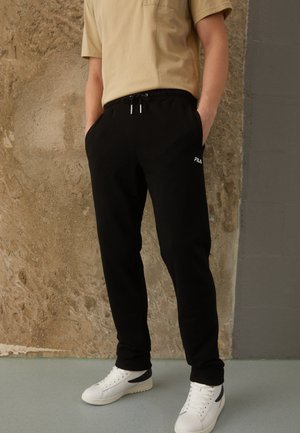 Pantaloni felpati neri con vita elasticizzata, tasche frontali e logo "FILA" bianco. Abbinati a una t-shirt beige e sneakers bianche.