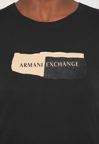 Tricou din bumbac negru, cu un design grafic care prezintă un bloc bej și negru, având inscripția "ARMANI EXCHANGE" în litere îndrăznețe.