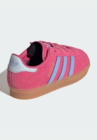 Sapatilha de camurça rosa com três riscas azul claro, um detalhe no calcanhar azul claro e uma sola de borracha de alcaçuz. "Gazelle" está bordado em amarelo.