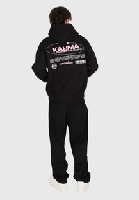 Personne portant un sweat à capuche noir avec le texte rose "KARMA" et un texte graphique blanc dans le dos, un pantalon noir et des chaussures blanches, se tenant devant un fond uni.