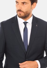 Completo blu strutturato con revers a punta, cravatta coordinata e camicia bianca; presenta un piccolo emblema a forma di forbice sul revers.