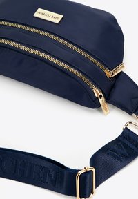 Námořnická modrá nylonová taška se dvěma zlatými zipovými přihrádkami, s obdélníkovou kovovou logem a texturovaným látkovým popruhem.