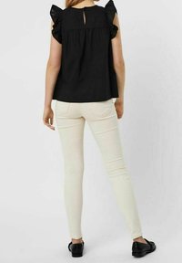Donna che indossa un top senza maniche nero con ruches e pantaloni skinny bianchi, con mocassini neri, in piedi di spalle su uno sfondo neutro.