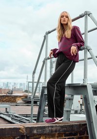 Top de manga larga a rayas en púrpura y rojo, pantalones negros con banda lateral blanca y calzado púrpura. La modelo posa sobre un andamio en la azotea.