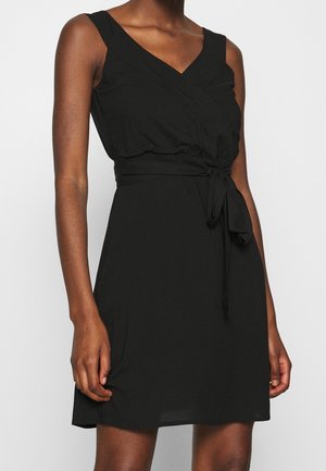 Ärmelloses schwarzes Wickelkleid mit V-Ausschnitt und Taillengürtel, getragen von einer Person mit brauner Haut vor einem schlichten Hintergrund.