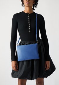 Borsa a tracolla blu in pelle con superficie texturizzata e hardware in tonalità argento; indossata con una blusa nera a coste e una gonna nera plissettata.