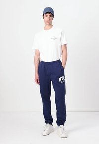 Balta T-krekls ar logo uz krūtīm, apvienots ar tumši ziliem sporta biksēm, uz kurām ir uzraksts "NYC". Komplektā ietilpst zila cepure un baltas sporta kurpes.