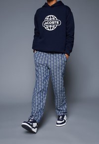 Sudadera con capucha azul marino con logo blanco de Lacoste, combinada con pantalones azules y blancos con un diseño geométrico, y zapatillas deportivas azules.