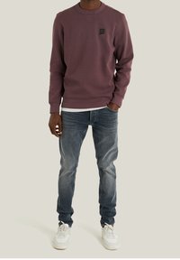 Maglione viola a coste con scollo rotondo, con una toppa con logo nero. Indossato sopra una camicia bianca e abbinato a jeans azzurri e scarpe da ginnastica bianche.