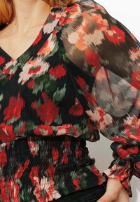 Blouse noire à motifs floraux rouges et verts, avec manches bouffantes transparentes et taille froncée. Tissu lisse et léger.