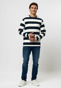 Man staat, draagt een zwart-wit gestreept shirt met lange mouwen, blauwe spijkerbroek en witte sneakers, met de handen samengevouwen voor zich.