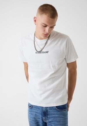 Jeune homme aux cheveux courts portant un t-shirt blanc « GUESS JEANS », un jean bleu, des chaînes argentées, regardant vers le bas sur un fond blanc uni.