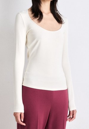 Kobieta ubrana w dopasowaną, długorękawową białą bluzkę z dekoltem w łódkę i luźne, wysokie spodnie w kolorze bordowym, stojąca na jednolitym tle.