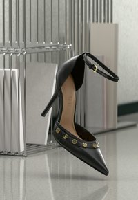 Crna kožna ženska cipela s visokim potpeticama i špicastim vrhom, nježni zlatni detalji na remenu i podesivi remen oko gležnja. Glatka tekstura, elegantan dizajn.