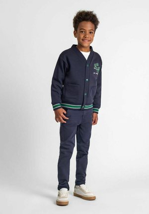 Marineblauer Cardigan mit grün-weißen gestreiften Saum, zwei vorderen Taschen und Knopfverschluss, kombiniert mit passenden marineblauen Hosen und weißen Turnschuhen.