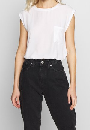 Vrouw draagt mouwloos wit shirt met een klein borstzakje en zwarte jeans met hoge taille, tegen een effen achtergrond.