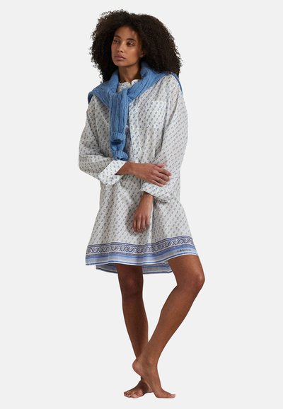 Femme aux cheveux bouclés portant une robe blanche à motifs floraux avec une bordure bleue et un pull bleu drapé sur ses épaules, debout pieds nus.