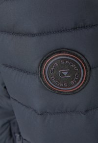 Mörkblå puffjacka med kviltad textur, med en rund, svart tygmärke som visar "CMS Sport" i röd skrift.