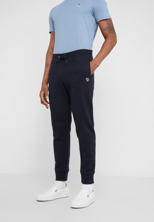 Hombre con camiseta azul claro, pantalones deportivos negros con cordón y pequeño logotipo bordado, calcetines blancos y zapatillas blancas tipo slip-on, de pie con la mano en el bolsillo.