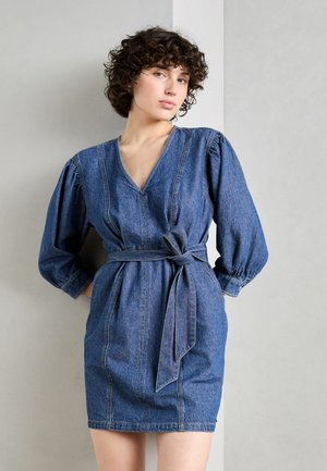 Abito in denim con scollo a V, maniche a sbuffo e cintura annodata in vita. Presenta una forma aderente e un colore blu medio con una texture sottile.