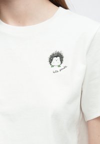 Biele bavlnené tričko s ilustráciou ježka so štruktúrovaným čiernym kožúškom, zelenými akcentmi a nápisom „kate spade“ pod ňou.