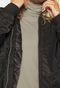 Giacca bomber nera con fodera trapuntata sopra un dolcevita grigio chiaro. Presenta una zip e polsini a coste. Tessuti morbidi e finitura liscia.