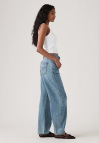 Γυναίκα με μακριά σγουρά μαλλιά φοράει λευκό αμάνικο τοπ, φαρδιά ανοιχτό μπλε τζιν παντελόνια και μαύρα διχτυωτά μπαλαρίνα παπούτσια, στέκεται πλάγια.