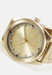 Reloj de acero inoxidable dorado con un acabado cepillado, marcadores de hora blancos y un diseño elegante. Presenta una corona y una pulsera de eslabones de metal.