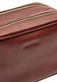 Bruine leren tas met twee gouden ritsen en een gegraveerde merknaam. Gestructureerd oppervlak met zichtbare stiksels. Rectangulaire vorm.