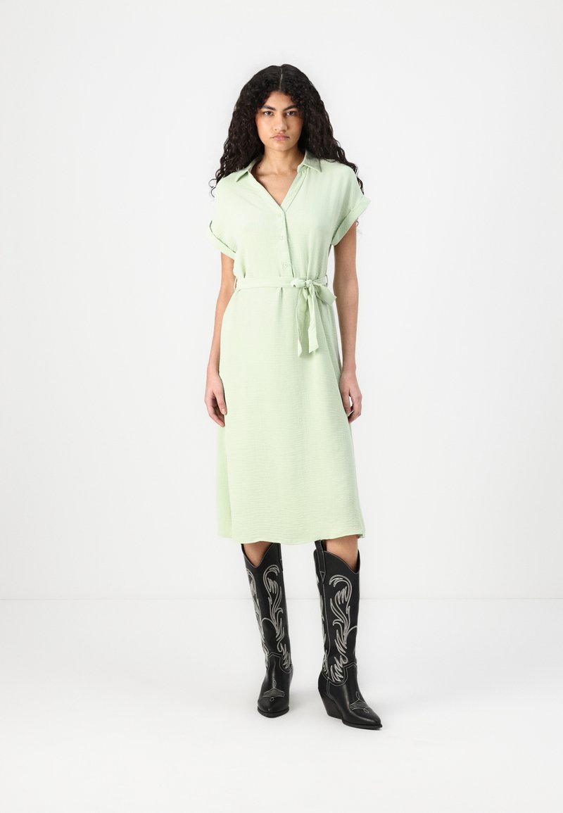 Rochie cămașă verde deschis cu o centură, mâneci scurte suflecate și deschidere cu nasturi. Cizme cowboy negre cu broderie argintie.