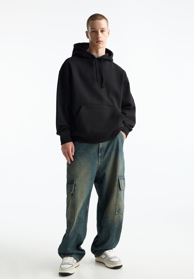 Schwarzer Hoodie mit Kängurutasche, kombiniert mit lockeren, ausgewaschenen blauen Cargo-Hosen mit Klapptaschen. Weiße Sneaker runden den Look ab.