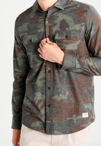 Chemise à manches longues avec un motif camouflage vert et marron, dotée de deux poches poitrine, d'une fermeture à boutons et d'un tissu à texture douce.