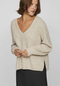 Světle béžový pletený svetr s výstřihem do V, žebrovanými dlouhými rukávy a bočními rozparky, který se vyznačuje měkkou texturou a uvolněným střihem.
