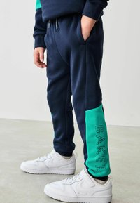 Marineblå sweatpants med teal farvede sidepaneler, prydet med ordet "BAKER" trykt vertikalt. Materialet er blødt, med elastisk talje og sidelommer.