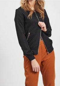 Vrouw in een zwarte ritsjack met gouden ritsen en brandoranje broek, hand die de rits van het jack halverwege naar beneden trekt.