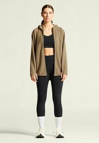 Bruine fleece jack met rits en capuchon, gedragen over een zwarte crop top en zwarte leggings. Witte sokken en sneakers maken de outfit compleet.