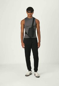 Borsa nera a tracolla indossata sopra una canottiera grigia, abbinata a pantaloni neri e sneaker bianche con dettagli neri. Materiale liscio e design minimalista.