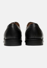 Scarpe in pelle nera con una texture liscia, punta arrotondata e dettagli brogue sulla parte superiore. Suola nera robusta con un design minimal.