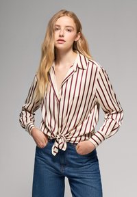 Bluzka z długim rękawem w paseczki w kolorze białym i burgundowym, związana w talii; w połączeniu z ciemnoniebieskimi dżinsami. Gładka tekstura materiału.