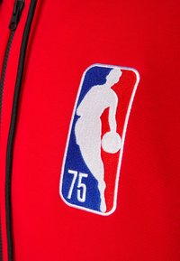 Tissu de sport rouge avec un logo NBA brodé en blanc, bleu et rouge, avec le numéro "75" en dessous. Détail de fermeture éclair visible.