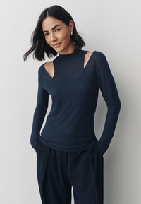 Top a maniche lunghe blu navy con collo alto finto, design con spalle scoperte e tessuto liscio. Abbinato a pantaloni scuri e ampi.