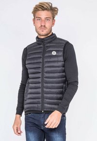 Veste rembourrée noire légère avec des coutures horizontales, fermeture éclair, col montant et poches latérales. Portée par-dessus une chemise noire à manches longues.